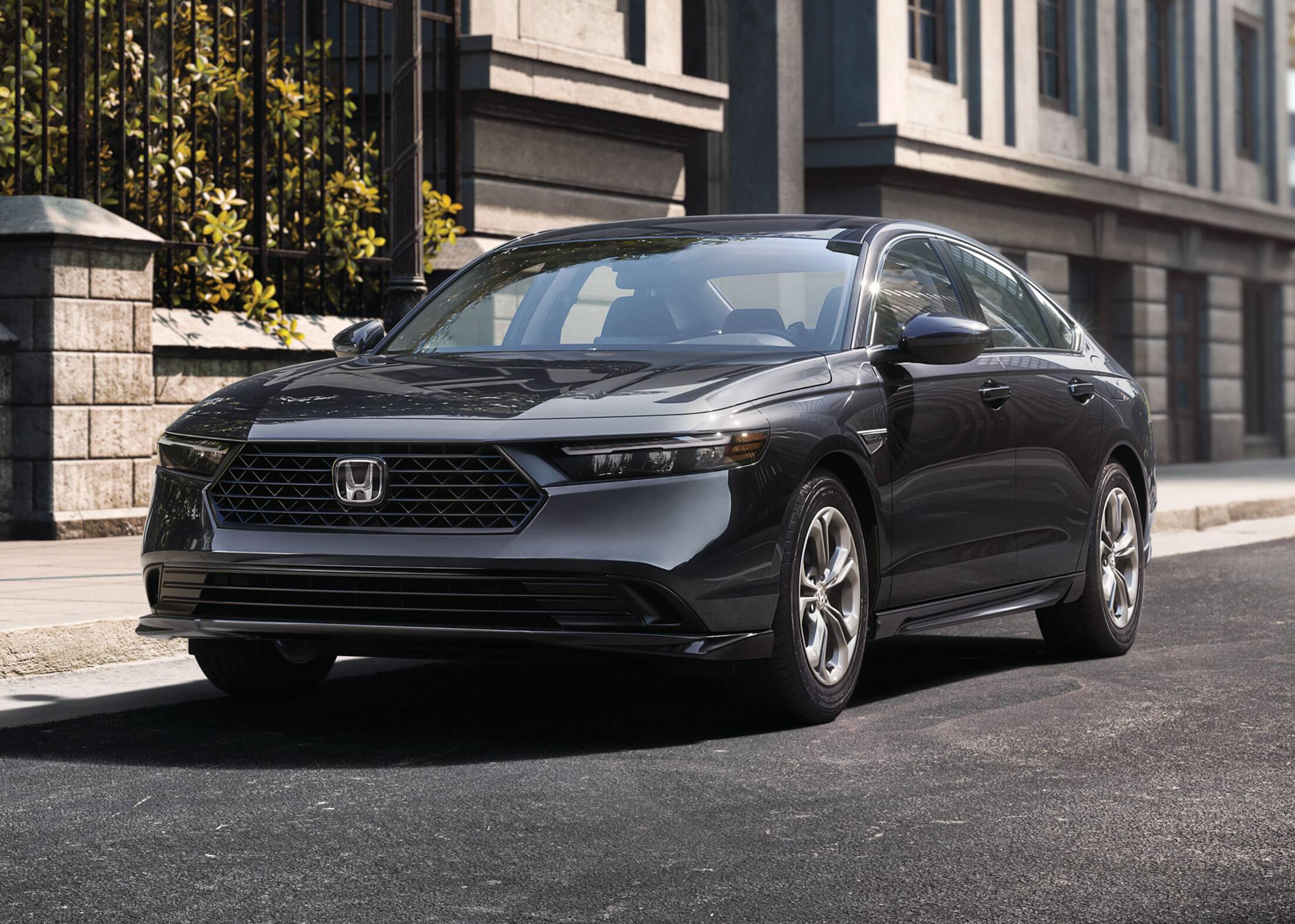 7/8 front driver-side view of the 2023 Honda Accord LX in Meteorite Gray Metallic, shown parked on the street next to a building and a city park with trees.