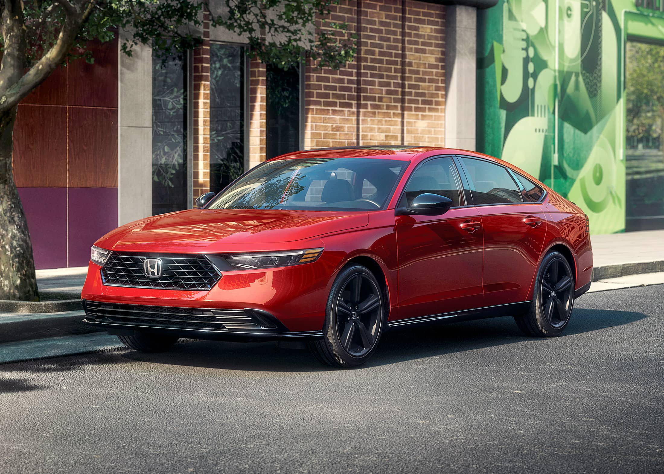 3/4 front driver-side view of the 2023 Honda Accord Sport-L Hybrid in Radiant Red Metallic, shown parked in front of an urban building with a colorful mural.