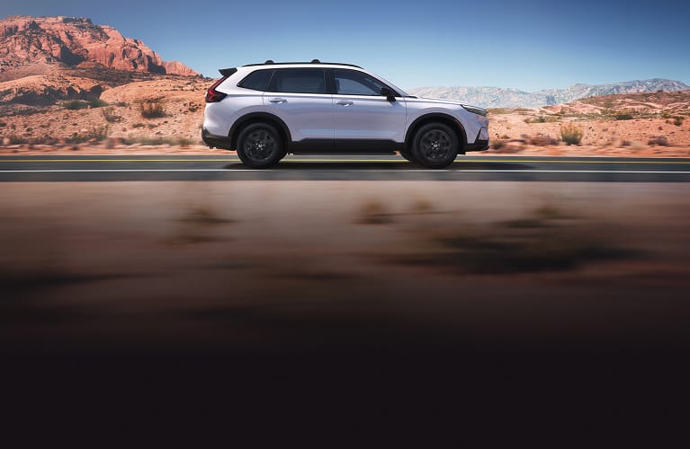 Perfil de la Honda CR-V Sport Touring Hybrid 2026 en Platinum White Pearl conduciendo por una pintoresca carretera desértica (SUV crossover compacta).
