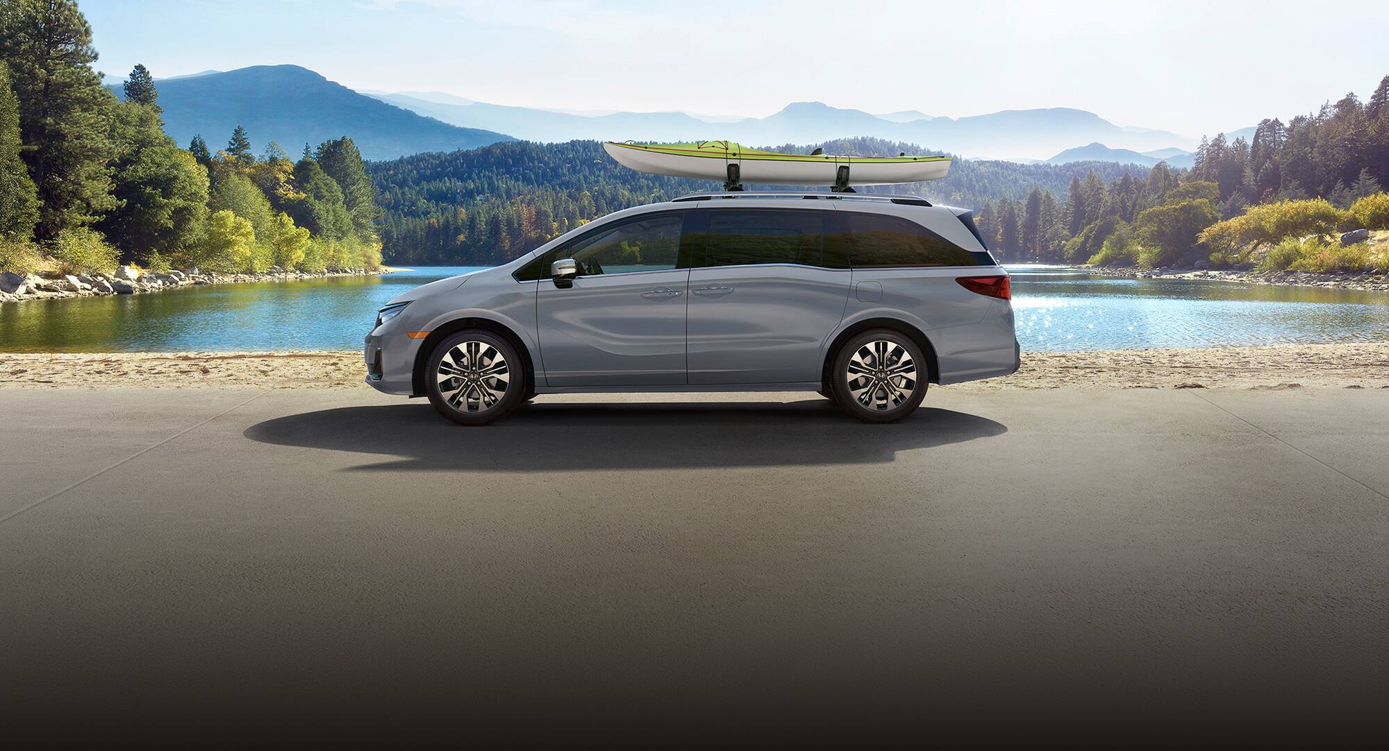 Perfil de la Honda Odyssey Elite 2027 en Sonic Gray Pearl estacionada junto a un lago con un kayak en el techo (minivan).